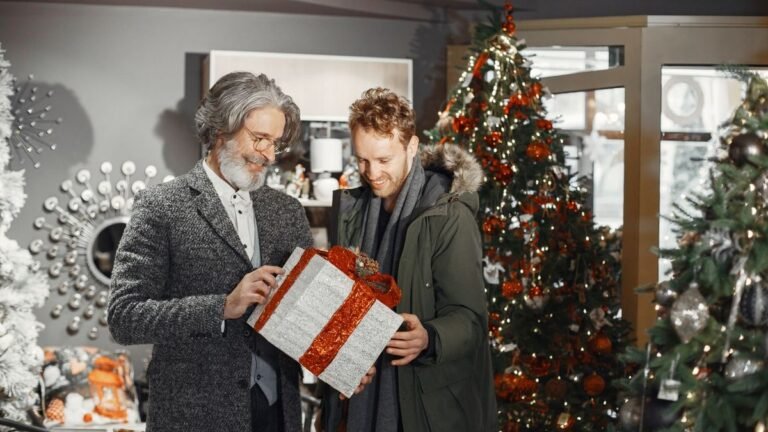 Two men smiling as they exchange a large wrapped Christmas gift beside a decorated tree