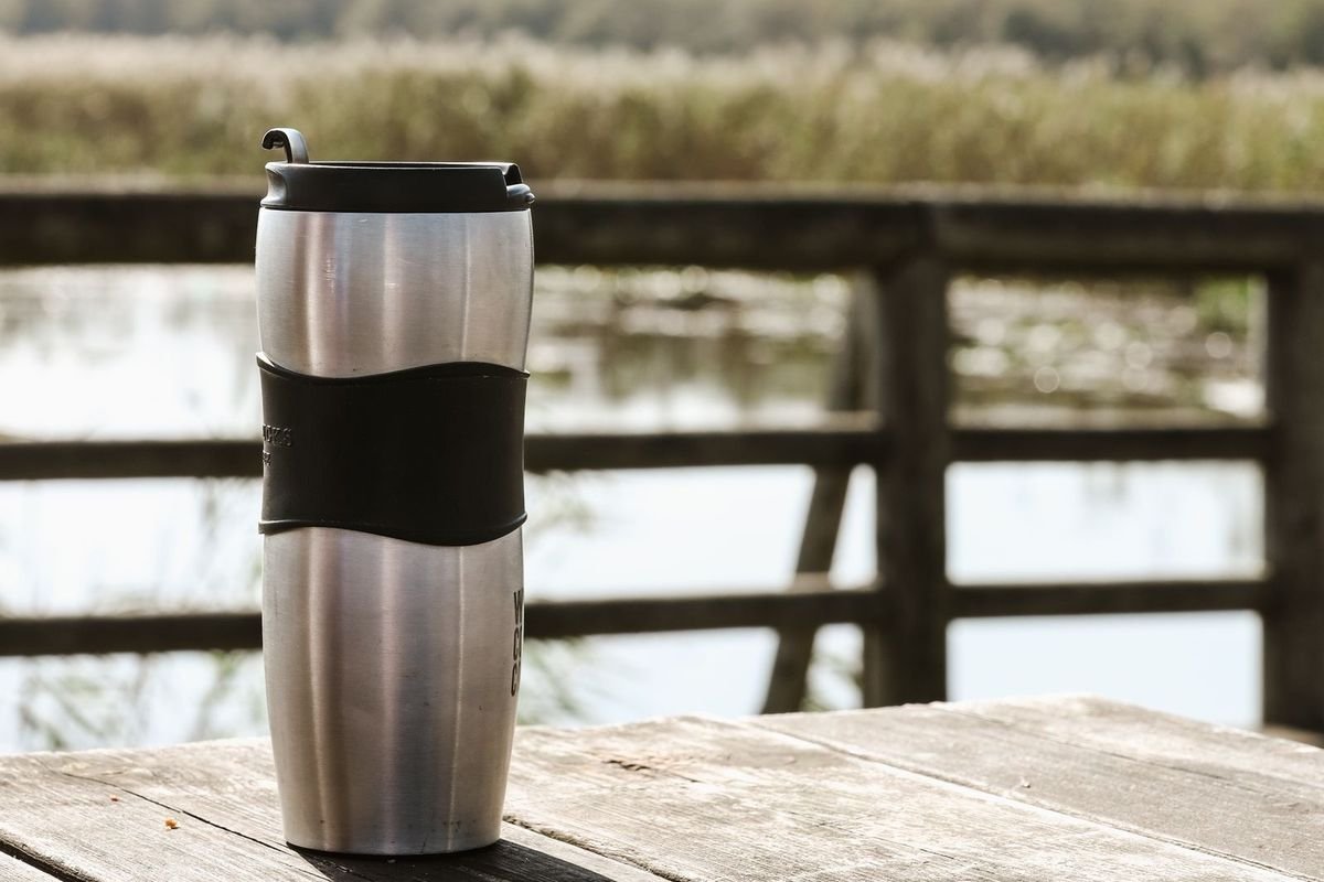 Stainless steel insulated travel mug on a wooden table by a lake (representative photo, not the exact product)
