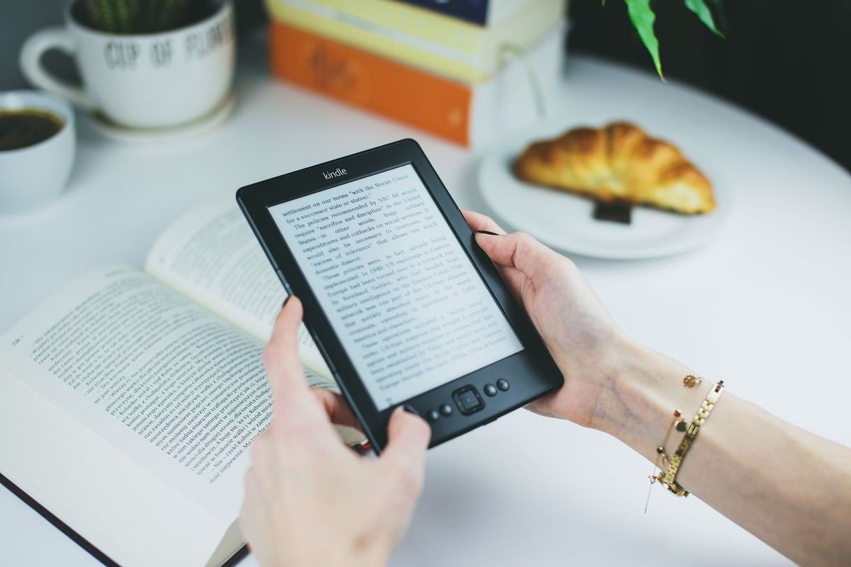 Hands holding an e-reader at a table with an open book and a croissant (representative photo, not the exact product)