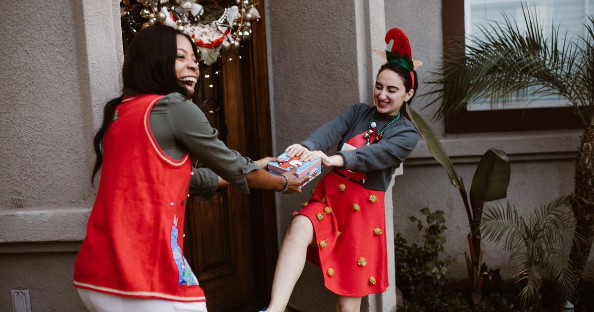 Two women laughing as they exchange a wrapped Christmas gift outside a front door