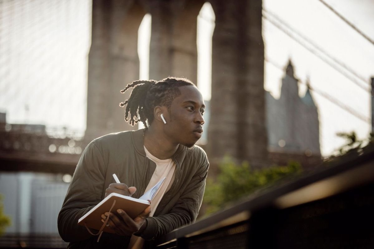 Man with wireless earbuds writing in a notebook near a city bridge