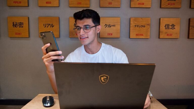Smiling man holding a smartphone at his desk with a laptop and mouse